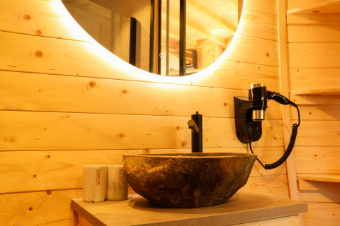 Baño moderno con paneles de madera, lavabo de piedra, espejo redondo y secador en Wellnest de Camping Vinkenhof.