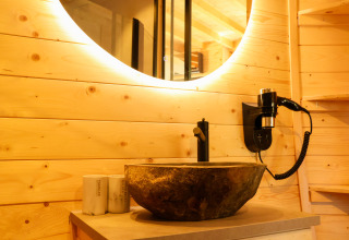 Baño moderno con paneles de madera, lavabo de piedra, espejo redondo y secador en Wellnest de Camping Vinkenhof.