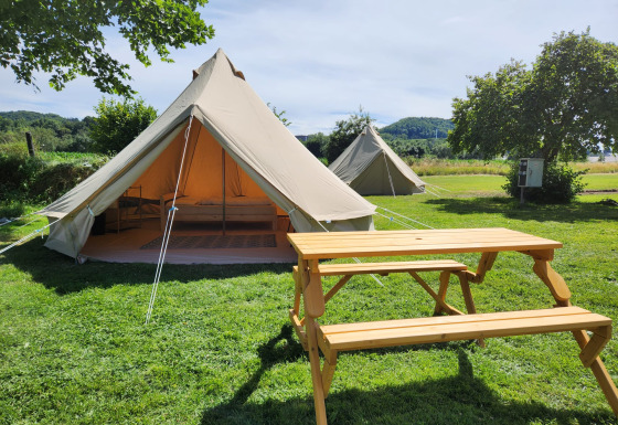 Safari tent Belltent op een groene camping met houten picknickbank, open tent en uitzicht op natuur.