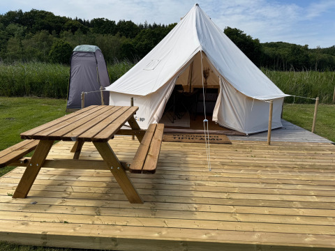 Tipi telt med træterrasse, picnicbord og telt i baggrunden omgivet af grønt ved en skov.