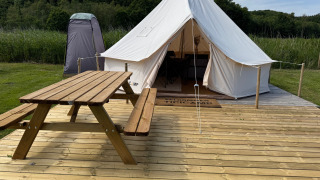 Tente tipi sur terrasse en bois avec table de pique-nique, toilette de campagne et verdure alentours.