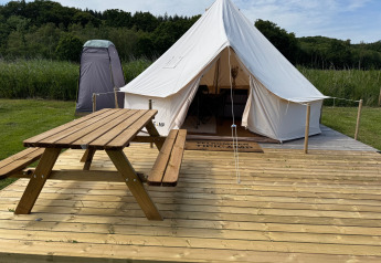 Tienda tipi sobre una tarima de madera con mesa de picnic, baño portátil y naturaleza verde de fondo.