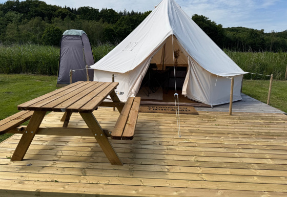 Tente tipi sur terrasse en bois avec table de pique-nique, toilette de campagne et verdure alentours.