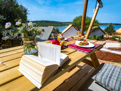 Colazione all'aperto su tavolo di legno con libro aperto, fiori e vista glamping ad Arena One 99, Croazia.