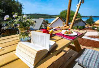 Desayuno al aire libre con libro abierto y flores en mesa de madera con vistas a glamping Arena One 99.