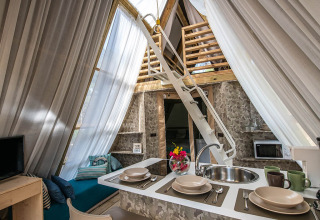 Interno accogliente di tenda safari con cucina, tavolo apparecchiato, scala per soppalco e tende chiare.
