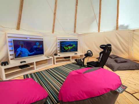 Area gaming in tenda con due TV, sedia da corsa Playseat, pouf colorati e console su pavimento in legno.