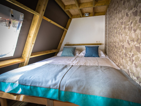 Cama acogedora con cojines azules dentro de una tienda safari con vigas de madera y papel tapiz militar.