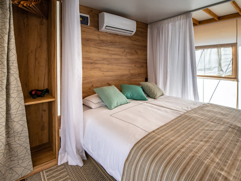 Chambre de tente safari moderne avec lit double, décoration en bois, rideaux blancs et climatisation.