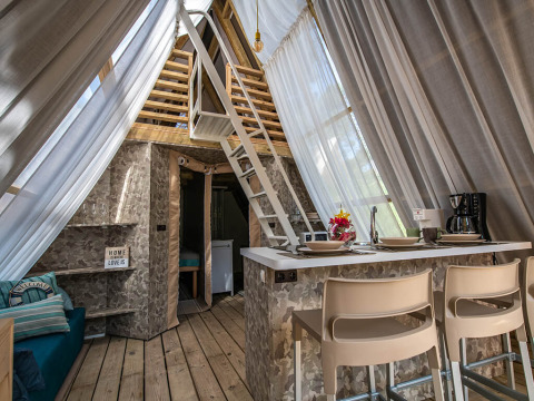 Intérieur d'une tente safari avec déco moderne, coin cuisine, espace salon et mezzanine sous voilages clairs.