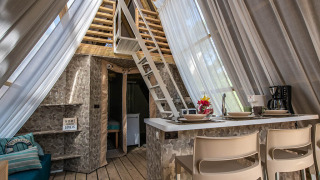 Binnenzicht van een safaritent met hedendaags interieur, keukentje, zithoek en een loft met lichte gordijnen.