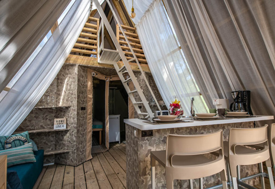 Binnenzicht van een safaritent met hedendaags interieur, keukentje, zithoek en een loft met lichte gordijnen.