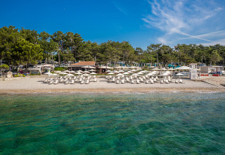 Strand met ligbedden en parasols bij Valamar Camping Ježevac, omgeven door bomen en helderblauw water.