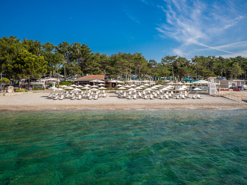 Strand met ligstoelen en parasols bij Valamar Camping Ježevac, helder blauw water en groene bomen erachter.