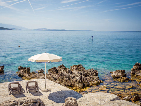 Loungebedden en parasol aan de rotsachtige kust bij Valamar Camping Ježevac, Kroatië, met helderblauwe zee.