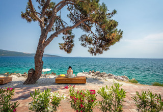 Vrouw zit op een bankje onder een boom met uitzicht op zee bij Valamar Camping Ježevac, Kroatië.