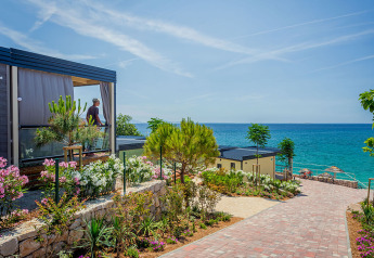 Lodge moderno con giardino fiorito e sentiero che porta alla splendida vista del mare e cielo azzurro.