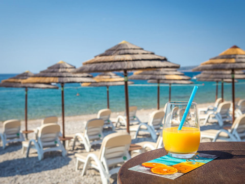 Glas sinaasappelsap op een tafel aan het strand bij Valamar Camping Ježevac in Kroatië, met ligstoelen en parasols.