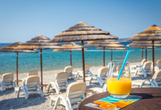 Glas sinaasappelsap op een tafel aan het strand bij Valamar Camping Ježevac in Kroatië, met ligstoelen en parasols.