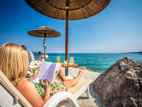 Femme se relaxant sur une chaise longue au bord de la mer, lisant un livre sous un parasol au Valamar Camping Ježevac.