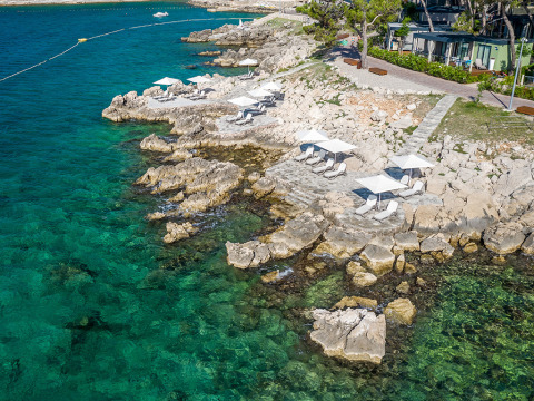 Luchtfoto van de rotsachtige kust bij Valamar Camping Ježevac met ligstoelen en parasols aan het heldere water.