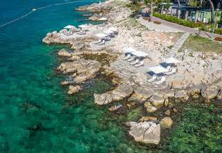 Luchtfoto van de rotsachtige kust bij Valamar Camping Ježevac met ligstoelen en parasols aan het heldere water.