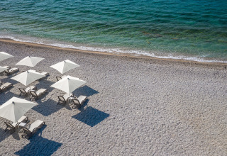 Solrige strandstole og hvide parasoller på stenstrand ved det klare hav ved Valamar Camping Ježevac, Kroatien.
