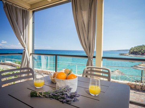 Terrasse ved havet med grå stole, juice, appelsiner og lavendel på bordet under et solrigt, blåt himmel.