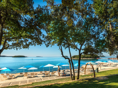 Strand von Valamar Camping Istra in Istrien, Kroatien, mit blauen Sonnenschirmen und Liegestühlen am Meer.