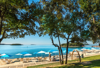 Strandbillede fra Valamar Camping Istra i Istrien, Kroatien, med blå parasoller, liggestole og havudsigt.