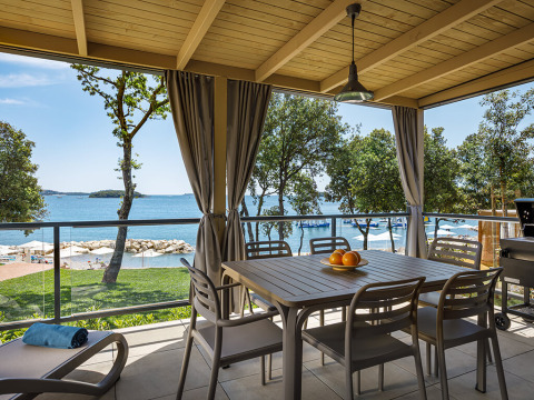 Overdekt lodge-terras met tafel en stoelen, met uitzicht op zee, omringd door bomen en een strand.