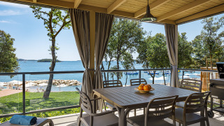 Overdekt lodge-terras met tafel en stoelen, met uitzicht op zee, omringd door bomen en een strand.