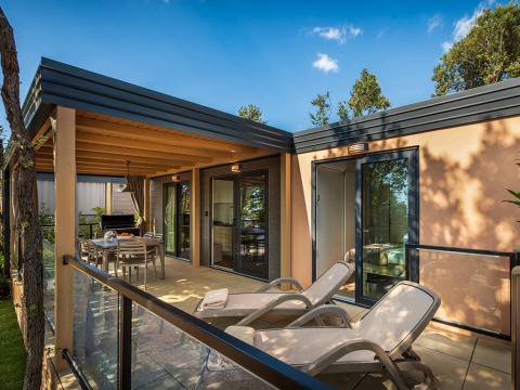 Buitenterras bij een lodge met ligstoelen, eethoek en glazen balustrade, omgeven door bomen.