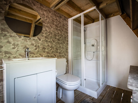 Baño en tienda safari con techo de madera, cabina de ducha, inodoro, lavabo y espejo redondo en la pared.