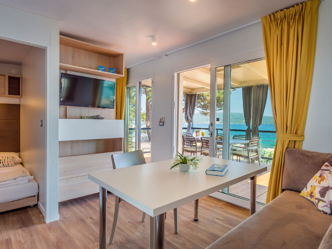 Moderne lodge met eettafel, bank, gele gordijnen en uitzicht op zee via grote glazen deuren naar terras.