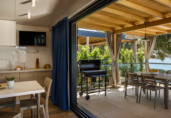 Moderne indendørs spisekøkken med udendørs terrasse, grill og havudsigt, omgivet af grønne planter.