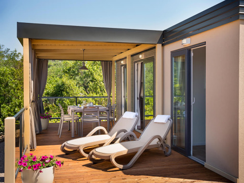 Zonnig lodge-terras met ligstoelen, een eettafel, bloemenpotten en groene natuur op de achtergrond.