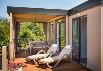 Terrasse ensoleillée de lodge avec chaises longues, table à manger, pots de fleurs et verdure en arrière-plan.
