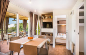 Licht lodge-interieur met eetruimte, slaapkamer, terras en grote ramen met zicht op de natuur.