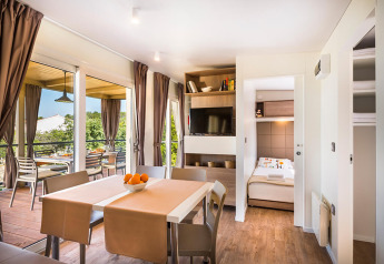 Licht lodge-interieur met eetruimte, slaapkamer, terras en grote ramen met zicht op de natuur.