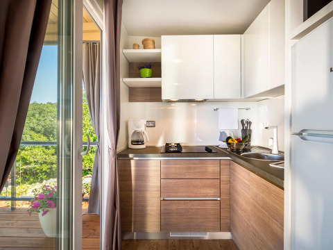 Moderne keuken in een lodge met houten kasten, witte planken en uitzicht op het groen buiten via de deur.