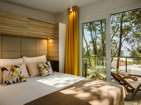 Moderne lodge slaapkamer met sierkussens, gele gordijnen en balkon met uitzicht op bomen en zee.