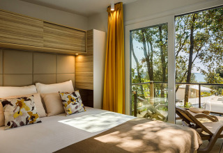 Moderne lodge slaapkamer met sierkussens, gele gordijnen en balkon met uitzicht op bomen en zee.