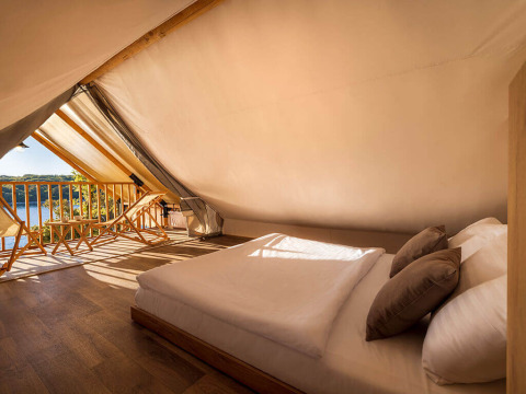 Interieur van een safaritent met tweepersoonsbed, houten stoelen en uitzicht op het meer in zonlicht.