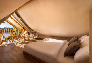 Interieur van een safaritent met tweepersoonsbed, houten stoelen en uitzicht op het meer in zonlicht.
