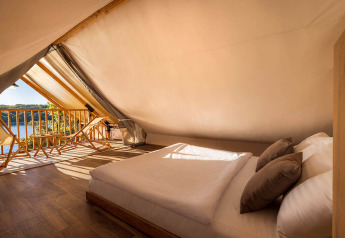Interior de una tienda safari con cama doble, sillas de madera y vista al lago bajo la luz del sol.