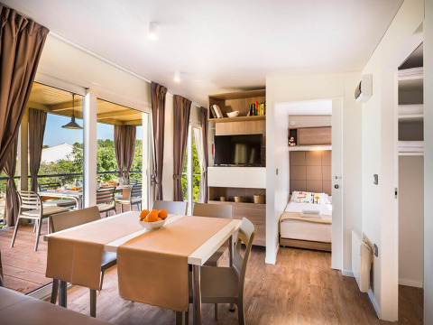 Moderne lodge woonkamer met eettafel, zicht op terras, grote ramen, tv en toegang tot slaapkamer.