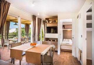 Moderne lodge leefruimte met eettafel, zicht op het terras, grote ramen, tv en toegang tot slaapkamer.