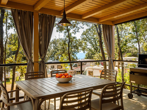 Overdekt terras bij een lodge met eettafel, stoelen, gordijnen en uitzicht op caravans in een groene omgeving.