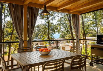 Terrasse couverte d’un lodge avec table à manger, chaises, rideaux et vue sur caravanes en pleine nature.